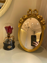 Oval gold decorative mirror with bow and floral detailing. A vintage-style accent piece perfect for vanity or tabletop decor.